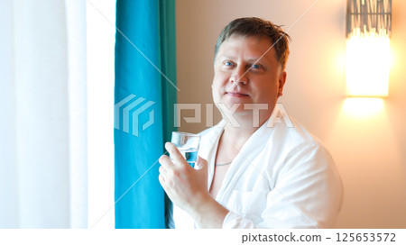 A man starts his morning with a glass of clean drinking water standing near the window A man starts his morning with a glass of clean drinking water standing near the window 125653572
