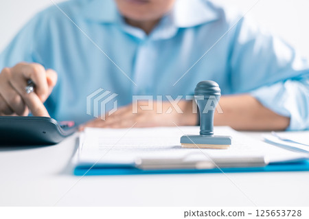 Notary placed their stamp of approval on the document as the lawyer and business person finalized the contract, ensuring all legal paperwork was in order. stamp, approval, document, contract, person. Notary placed their stamp of approval on the document as the lawyer and business person finalized the contract, ensuring all legal paperwork was in order. stamp, approval, document, contract, person. 125653728