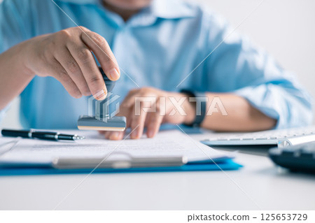 Notary placed their stamp of approval on the document as the lawyer and business person finalized the contract, ensuring all legal paperwork was in order. stamp, approval, document, contract, person. 125653729