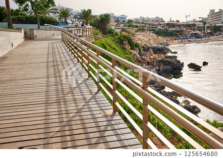 A wooden boardwalk with metal railing leads along the coast, offering a scenic view of the sea. A wooden boardwalk with metal railing leads along the coast, offering a scenic view of the sea. 125654680