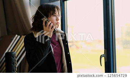 A girl speaks on her phone while standing near a bright window, her expression calm and focused. The photo reflects themes of technology, connection, and real-time interaction. A girl speaks on her phone while standing near a bright window, her expression calm and focused. The photo reflects themes of technology, connection, and real-time interaction. 125654729