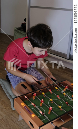 A boy in a red shirt smiles while playing foosball, enjoying a fun and competitive moment at home. 125654751