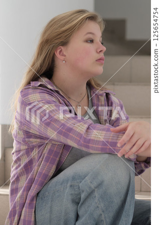 teenage girl rests on the steps with a pensive expression, dressed casually in a plaid shirt and jeans. This photo highlights introspection and the beauty of simplicity. 125654754