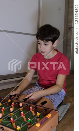 A boy in a red shirt smiles while playing foosball, enjoying a fun and competitive moment at home. 125654803