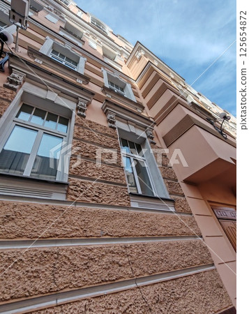 A low-angle view of a historic peach facade with ornate white window designs and a prominent surveillance camera. The image emphasizes the coexistence of traditional architecture and contemporary A low-angle view of a historic peach facade with ornate white window designs and a prominent surveillance camera. The image emphasizes the coexistence of traditional architecture and contemporary 125654872