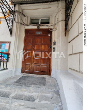 A sturdy wooden door with square panels, surrounded by an aged white facade, stone steps, and wrought iron canopy supports. The image highlights urban entrances and vintage architectural charm. A sturdy wooden door with square panels, surrounded by an aged white facade, stone steps, and wrought iron canopy supports. The image highlights urban entrances and vintage architectural charm. 125654884