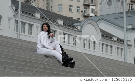 A woman in a white tracksuit sits on outdoor stairs with kangaroo boots resting beside her, taking a break from exercise. The contrast between historical architecture and modern fitness gear reflects 125655150