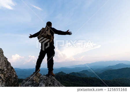Male tourist on top of rocky mountain. A peaceful man meditating yoga relaxing alone standing and spreading arms on a mountain top at sunrise with nature landscape. Male tourist on top of rocky mountain. A peaceful man meditating yoga relaxing alone standing and spreading arms on a mountain top at sunrise with nature landscape. 125655168
