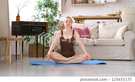 Seated on a blue yoga mat, a woman in a brown outfit closes her eyes in a tranquil living room setting. This photo captures the essence of home-based meditation and its role in stress reduction. Seated on a blue yoga mat, a woman in a brown outfit closes her eyes in a tranquil living room setting. This photo captures the essence of home-based meditation and its role in stress reduction. 125655175