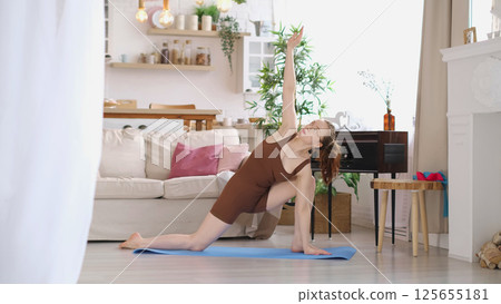 Performing a yoga stretch, a woman in a brown tank top and shorts balances on a blue mat in a well-lit living room. This photo captures the essence of home fitness and the pursuit of physical well 125655181