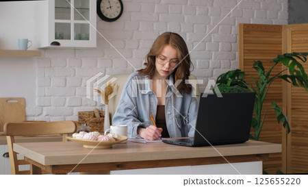 Seated at a kitchen table, a woman in glasses writes in a notebook while using a laptop. This photo captures the essence of modern remote work and the importance of a conducive workspace at home. 125655220