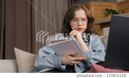 A woman in a denim jacket sits on a couch, focused and thoughtful, holding a notebook and pen while looking at her laptop. This image captures the intensity and concentration of remote work or study 125655226