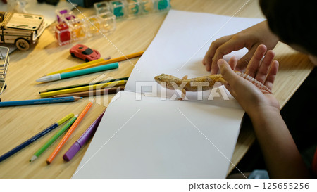 A child interacts with a leopard gecko while preparing to draw on a blank page. The image highlights the integration of creativity and responsibility in pet ownership. A child interacts with a leopard gecko while preparing to draw on a blank page. The image highlights the integration of creativity and responsibility in pet ownership. 125655256