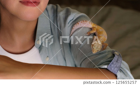 A gecko perched on a woman s shoulder looks outward, embodying curiosity and calm. The image connects with themes of exotic wildlife and respectful handling of unique creatures. A gecko perched on a woman s shoulder looks outward, embodying curiosity and calm. The image connects with themes of exotic wildlife and respectful handling of unique creatures. 125655257