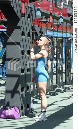 A woman in a blue sports outfit takes a break to hydrate during her workout at an outdoor gym, highlighting the importance of staying hydrated during exercise. This image emphasizes the balance A woman in a blue sports outfit takes a break to hydrate during her workout at an outdoor gym, highlighting the importance of staying hydrated during exercise. This image emphasizes the balance 125655264