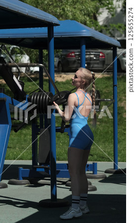 A woman in a blue bodysuit adjusts weights on exercise equipment at an outdoor gym, emphasizing her focus on building strength and endurance. This image connects the concept of outdoor workouts with 125655274