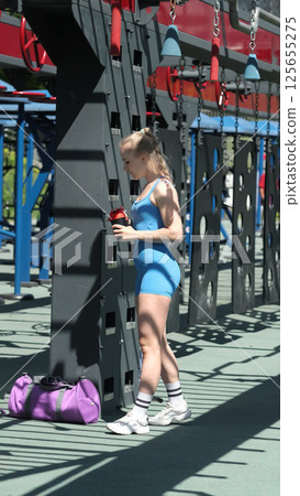 A woman in blue sportswear rehydrates with a water bottle during her outdoor workout session, standing by gym equipment. The photo showcases the crucial practice of drinking water to stay energized 125655275