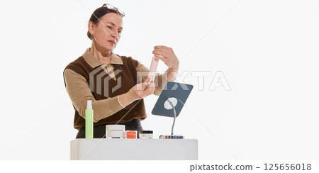Mature woman checking skincare product near mirror against white background. Concept of beauty routine, 125656018
