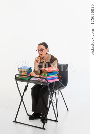 Older woman with pencil at lips, surrounded by books, contemplating deeply against white studio background. Older woman with pencil at lips, surrounded by books, contemplating deeply against white studio background. 125656059