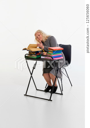 Overloaded with study. Elderly woman at desk surrounded by colorful books against white studio background, deeply focused on reading, multitasking study. Overloaded with study. Elderly woman at desk surrounded by colorful books against white studio background, deeply focused on reading, multitasking study. 125656060