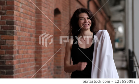 A smiling woman in sportswear walks along a brick wall, holding her jacket over her shoulder. The combination of urban architecture and active lifestyle reflects modern wellness trends. 125656092