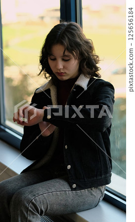 A young girl sitting by a window checks her watch with a thoughtful expression. This image highlights themes of time, mindfulness, and youth in a calm, natural light setting. 125656184