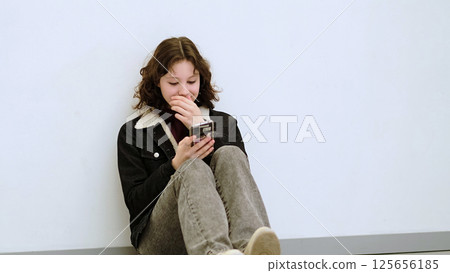 A young girl sits on the floor smiling as she looks at her smartphone. This image highlights themes of digital connection, enjoyment, and the lighthearted moments technology can bring. A young girl sits on the floor smiling as she looks at her smartphone. This image highlights themes of digital connection, enjoyment, and the lighthearted moments technology can bring. 125656185