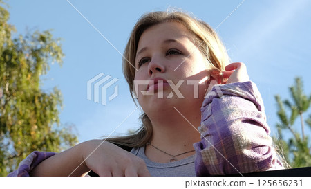 A blonde girl with tears in her eyes looks thoughtfully away, surrounded by a clear sky and trees. This image highlights themes of emotional depth and personal reflection. 125656231