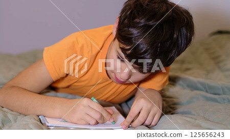 A young boy uses a crayon to draw in a notebook while lying on a comfortable bed. The setting highlights a peaceful indoor activity and the power of self-expression. A young boy uses a crayon to draw in a notebook while lying on a comfortable bed. The setting highlights a peaceful indoor activity and the power of self-expression. 125656243