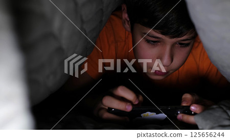 young boy uses his smartphone under a blanket, with the soft light creating a cozy yet mysterious atmosphere. The image reflects a blend of technology and youthful exploration. young boy uses his smartphone under a blanket, with the soft light creating a cozy yet mysterious atmosphere. The image reflects a blend of technology and youthful exploration. 125656244