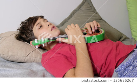 A young boy sits with a vibrant green toy guitar, showcasing his musical exploration and creative spirit. A young boy sits with a vibrant green toy guitar, showcasing his musical exploration and creative spirit. 125656267