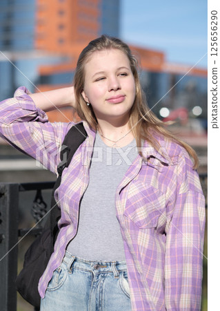 young woman closes her eyes and enjoys the sunlight with a content expression, standing near a modern cityscape. This image conveys peace and a connection to simple joys. 125656290