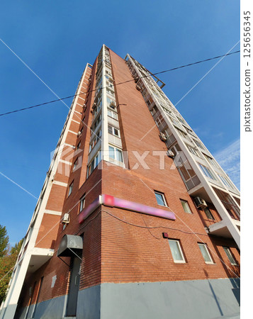 A tall red and beige brick residential building with multiple balconies, air conditioning units, and a sidewalk below. The structure reflects the typical architecture of urban apartment blocks in 125656345