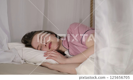 A young woman in pink sleepwear lies peacefully on a white pillow with sheer curtains around her, reflecting a serene and restful state. The scene emphasizes the importance of a comfortable sleeping A young woman in pink sleepwear lies peacefully on a white pillow with sheer curtains around her, reflecting a serene and restful state. The scene emphasizes the importance of a comfortable sleeping 125656705