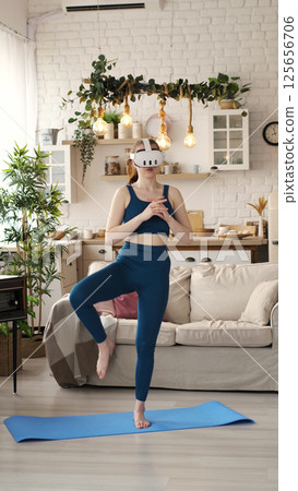A woman in a teal outfit practices a balancing pose while wearing a VR headset in her stylish living room. This image highlights the innovative use of virtual reality technology in home fitness A woman in a teal outfit practices a balancing pose while wearing a VR headset in her stylish living room. This image highlights the innovative use of virtual reality technology in home fitness 125656706