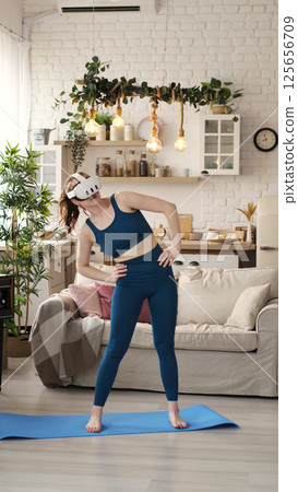 A woman in a teal outfit practices a balancing pose while wearing a VR headset in her stylish living room. This image highlights the innovative use of virtual reality technology in home fitness A woman in a teal outfit practices a balancing pose while wearing a VR headset in her stylish living room. This image highlights the innovative use of virtual reality technology in home fitness 125656709