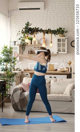 Engaged in a virtual reality workout, a woman in teal activewear balances on one foot in her modern living room. This photo captures the dynamic integration of VR technology with exercise. Engaged in a virtual reality workout, a woman in teal activewear balances on one foot in her modern living room. This photo captures the dynamic integration of VR technology with exercise. 125656710