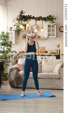 Balancing on one foot, a woman in teal workout clothes engages in a VR fitness session in her stylish living room. The photograph promotes the innovative blend of technology and exercise in Balancing on one foot, a woman in teal workout clothes engages in a VR fitness session in her stylish living room. The photograph promotes the innovative blend of technology and exercise in 125656713