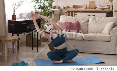 Engaged in a virtual reality workout, a woman in teal activewear stretches on a yoga mat in her stylish living room. This photo captures the innovative approach to home fitness using VR technology. Engaged in a virtual reality workout, a woman in teal activewear stretches on a yoga mat in her stylish living room. This photo captures the innovative approach to home fitness using VR technology. 125656715