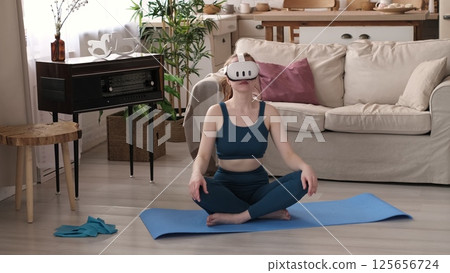 A woman in a teal outfit stretches while wearing a VR headset, seated on a blue yoga mat in her cozy living room. This image highlights the fusion of fitness and virtual reality technology in modern A woman in a teal outfit stretches while wearing a VR headset, seated on a blue yoga mat in her cozy living room. This image highlights the fusion of fitness and virtual reality technology in modern 125656724