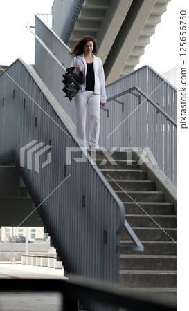 A woman dressed in sportswear moves down a metal staircase, carrying specialized jumping boots. The industrial backdrop emphasizes the fusion of fitness, architecture, and city movement. A woman dressed in sportswear moves down a metal staircase, carrying specialized jumping boots. The industrial backdrop emphasizes the fusion of fitness, architecture, and city movement. 125656750