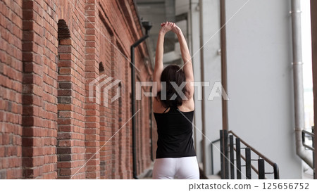 A woman in sportswear stretches her arms upward while standing near a brick wall. The blend of fitness and urban architecture symbolizes balance, movement, and well-being in modern city life. 125656752