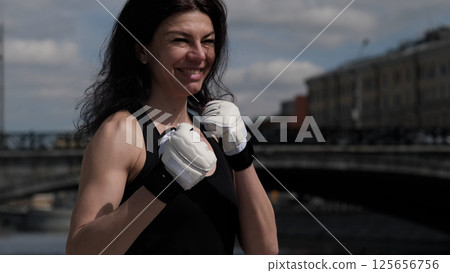 A smiling woman in boxing gloves stands confidently outdoors with a cityscape in the background. The growing trend of women in combat sports reflects empowerment, resilience, and self-confidence. 125656756