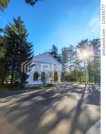 A bright neoclassical building surrounded by tall trees and illuminated by the morning sun. The soft shadows cast by the trees create a tranquil and serene atmosphere. 125656777
