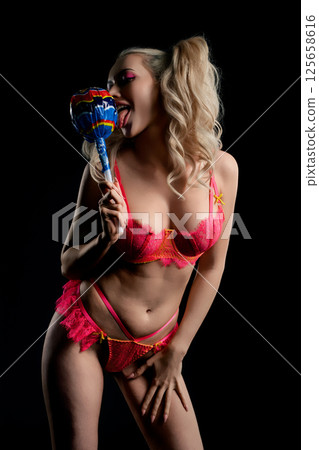 Woman in pink lingerie playfully poses with lollipop against a dark background 125658616