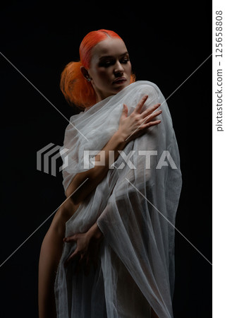 Red-haired woman poses nude with sheer fabric against dark background 125658808