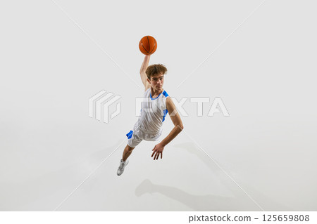 Aerial view photo of young male basketball athlete flying through air, arm raised to score final goal against white studio background. Aerial view photo of young male basketball athlete flying through air, arm raised to score final goal against white studio background. 125659808