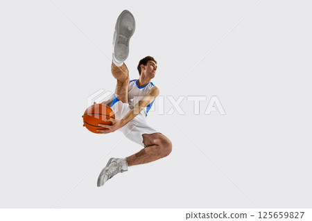 Low angle view. Athletic young man in white-blue game outfit sprinting with basketball, practicing dribbling technique with intensity against white background. 125659827