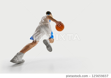 Low angle view. Young basketball player runs forward with determination, gripping ball tightly and dressed in game uniform against white background. Low angle view. Young basketball player runs forward with determination, gripping ball tightly and dressed in game uniform against white background. 125659837