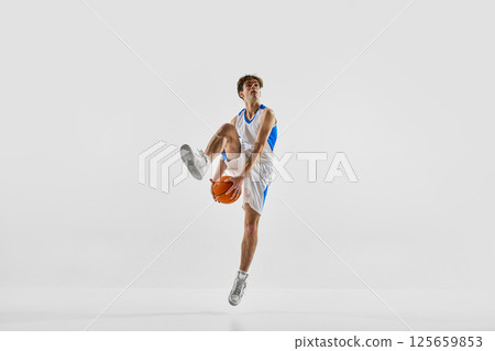 Focused basketball player jumping with raised leg holding ball against white studio background, showing intensity and power. Focused basketball player jumping with raised leg holding ball against white studio background, showing intensity and power. 125659853